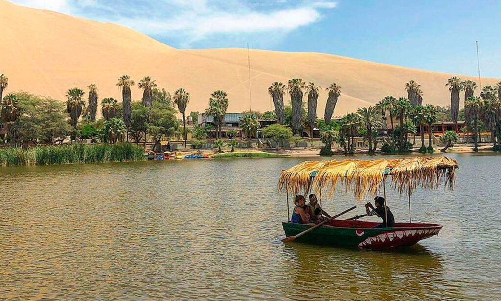 Bote con techo de palma navegando sobre la laguna de Huacachina, rodeada de palmeras y dunas