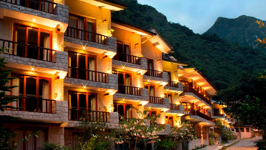 Fachada iluminada de un hotel en Aguas Calientes al anochecer, rodeado por el paisaje montañoso