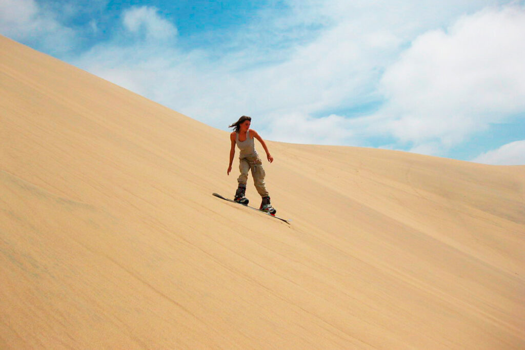 Mujer descendiendo de pie en tabla por una duna alta del desierto de Huacachina