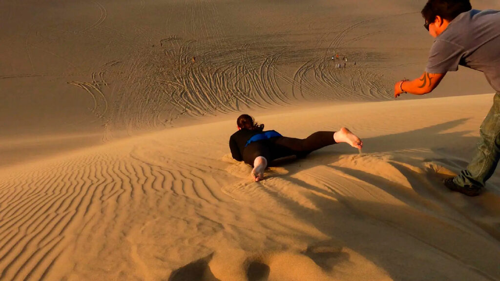 Instructor guiando a un visitante que practica sandboarding acostado en una duna suave de Huacachina
