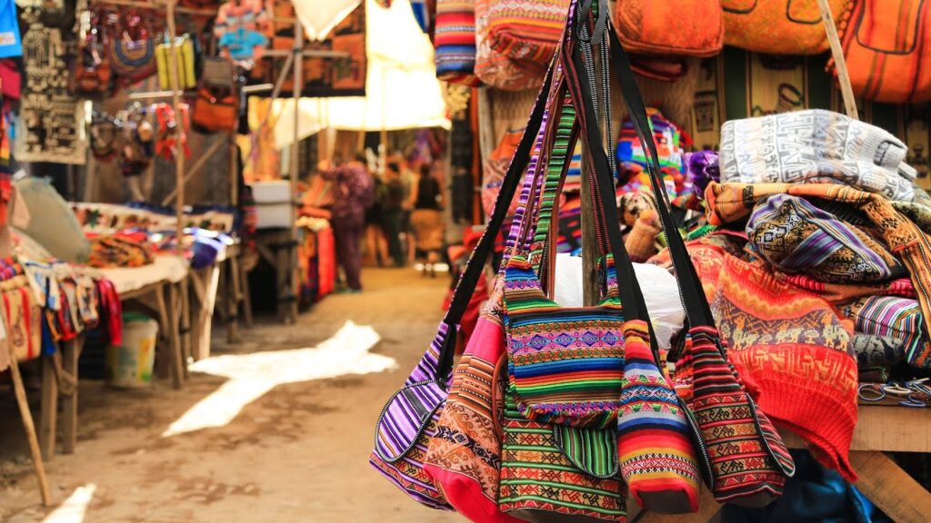 Puestos de artesanía andina en el mercado de Pisac, con textiles tradicionales y bolsos coloridos exhibidos en un pasillo del mercado
