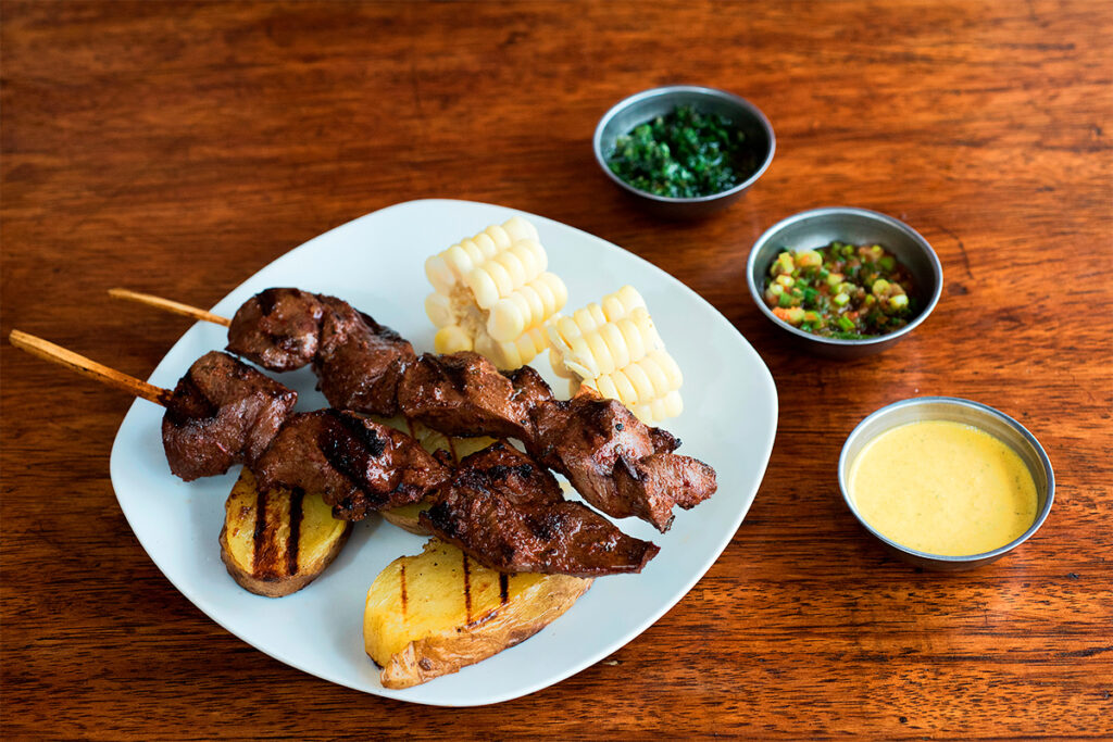 Plato de anticuchos de corazón de res con papa, choclo, ají de huacatay y salsas, sobre una mesa de madera