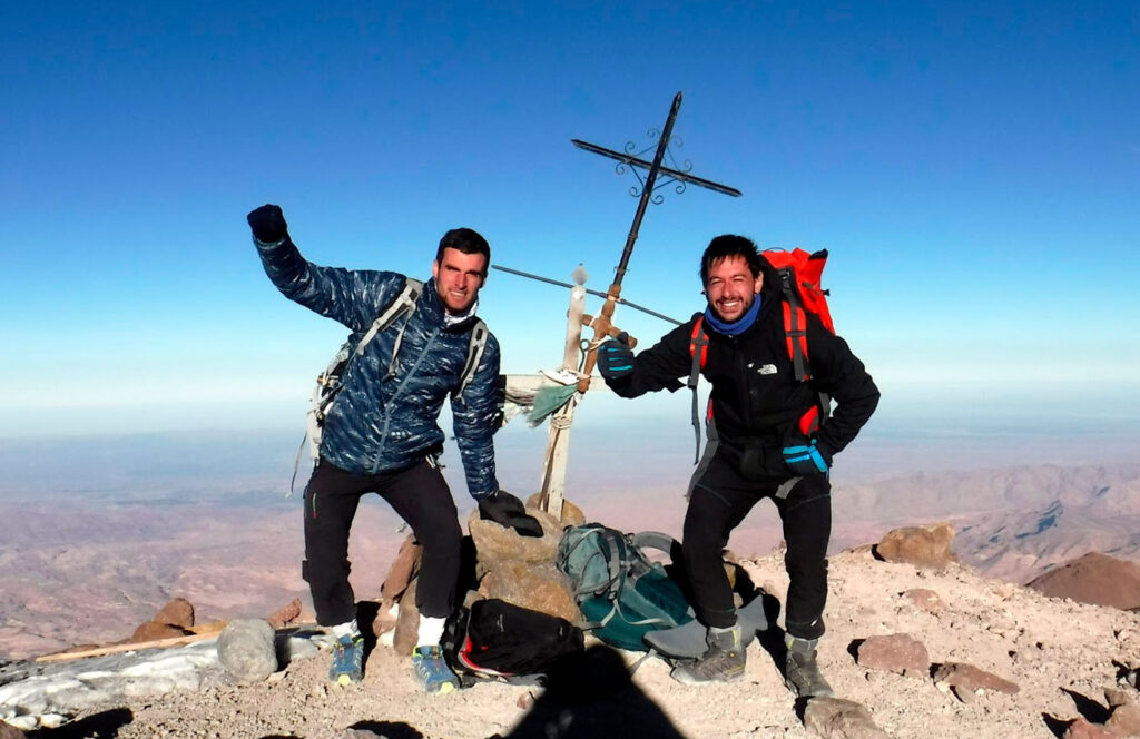 Dos montañistas celwebrando en la cima de un volcán del sur del perú, con mochilas, bastones y una cruz andina, bajo el cielo despejado