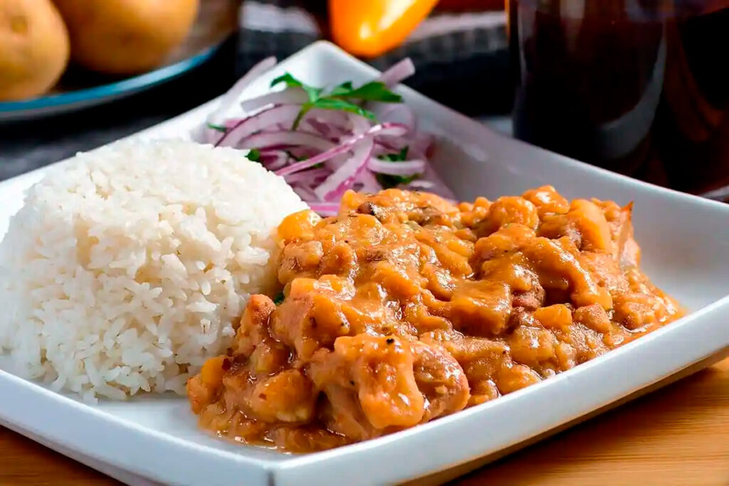 Carapulcra peruana servida con arroz vlanco y salsa criolla, en plato rectangular sobre una mesa de madera