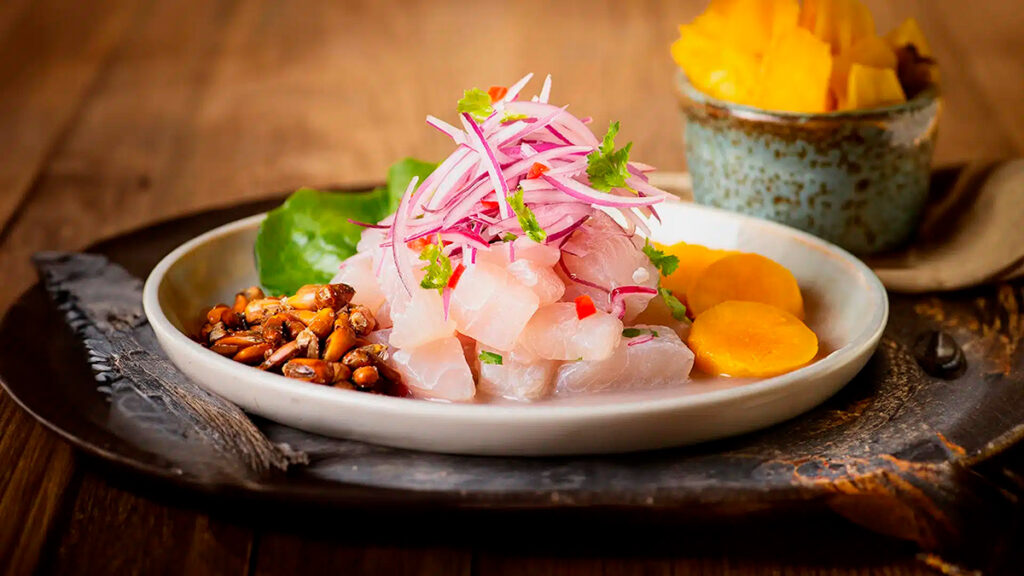 Plato de ceviche peruano decorado con camote, maíz tostado y cebolla morada, servido sobre una fuente de madera