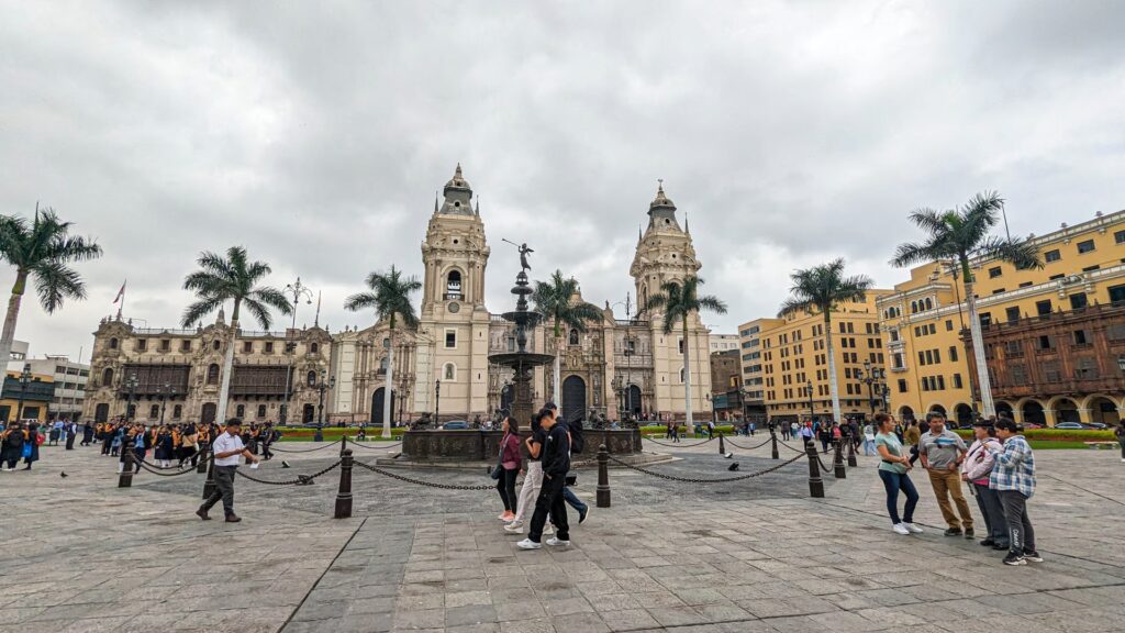 Vista panorámica de la Plaza Mayor de Lima con la Catedral y edificios coloniales en un día nublado, con personas transitando la plaza.