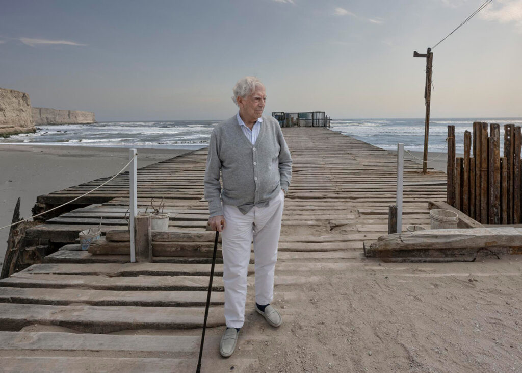 Mario Vargas Llosa de pie en un muelle de madera junto al mar, con bastón en mano, mirando hacia el horizonte.