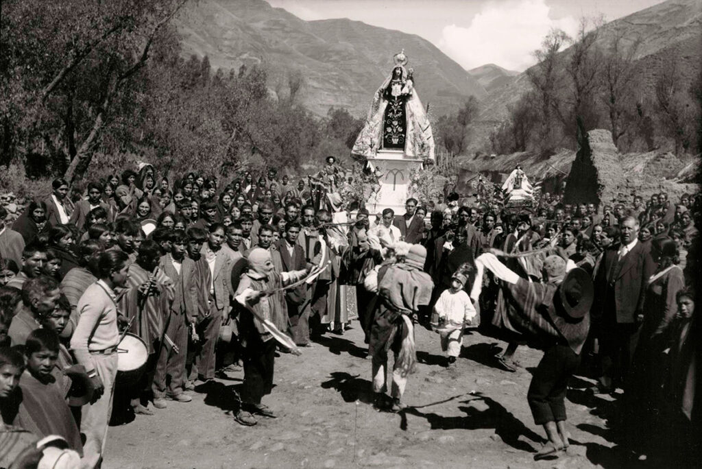 Fotografía antigua de una procesión en Andahuaylillas, con un santo en andas, rodeado por fieles y músicos, tomada por Martín Chambi