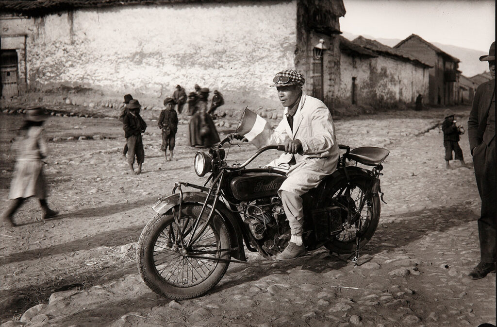 Fotografía antigua en blanco y negro de un motociclista en Cusco tomada por Martín Chambi, con fondo urbano y personas caminando alrededor 