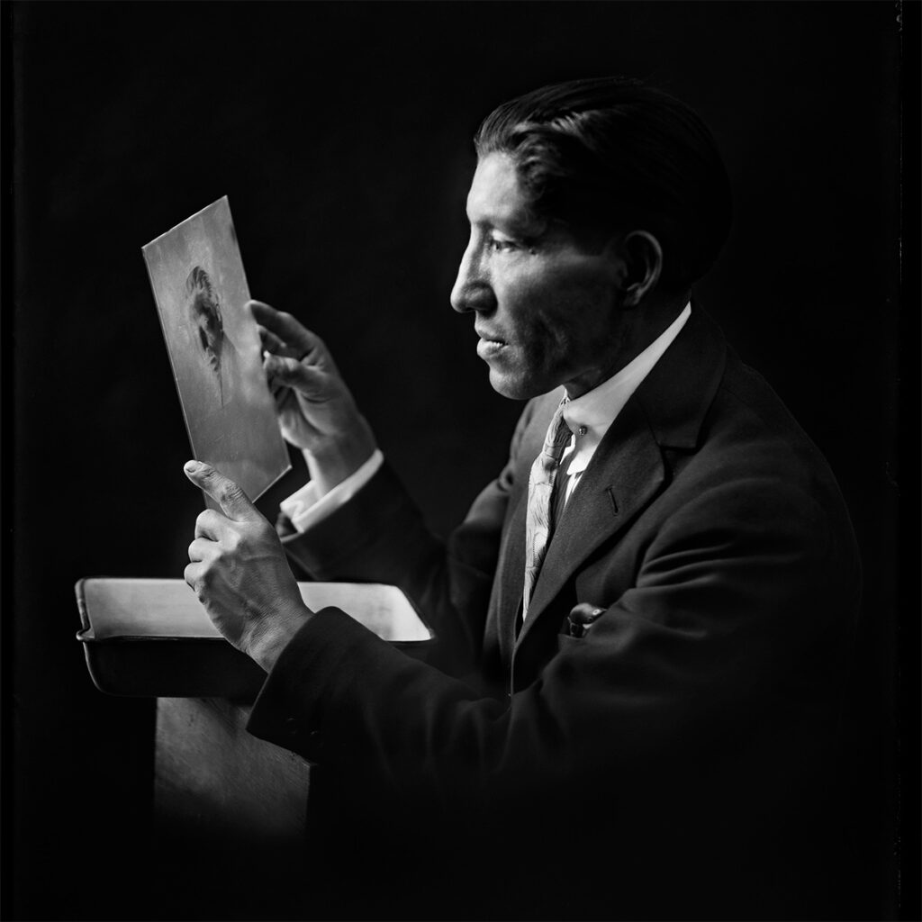 Retrato de Martín Chambi sosteniendo un negativo fotográfico, en blanco y negro, con fondo oscuro