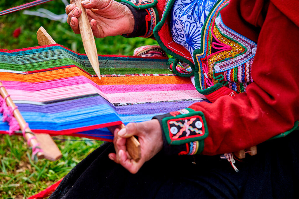 Manos de una tejedora andina de chinchero trabajando en un telar de cintura con hilos de colores vivos.