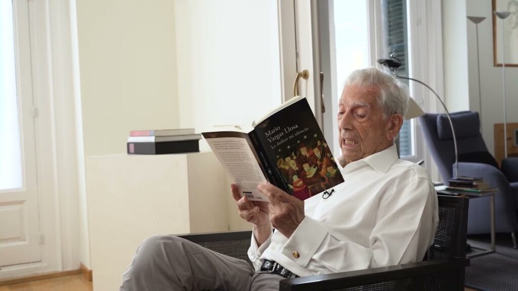 Mario Vargas Llosa sentado en un sillón, leyendo su libro Le dedico mi silencio en una habitación iluminada.
