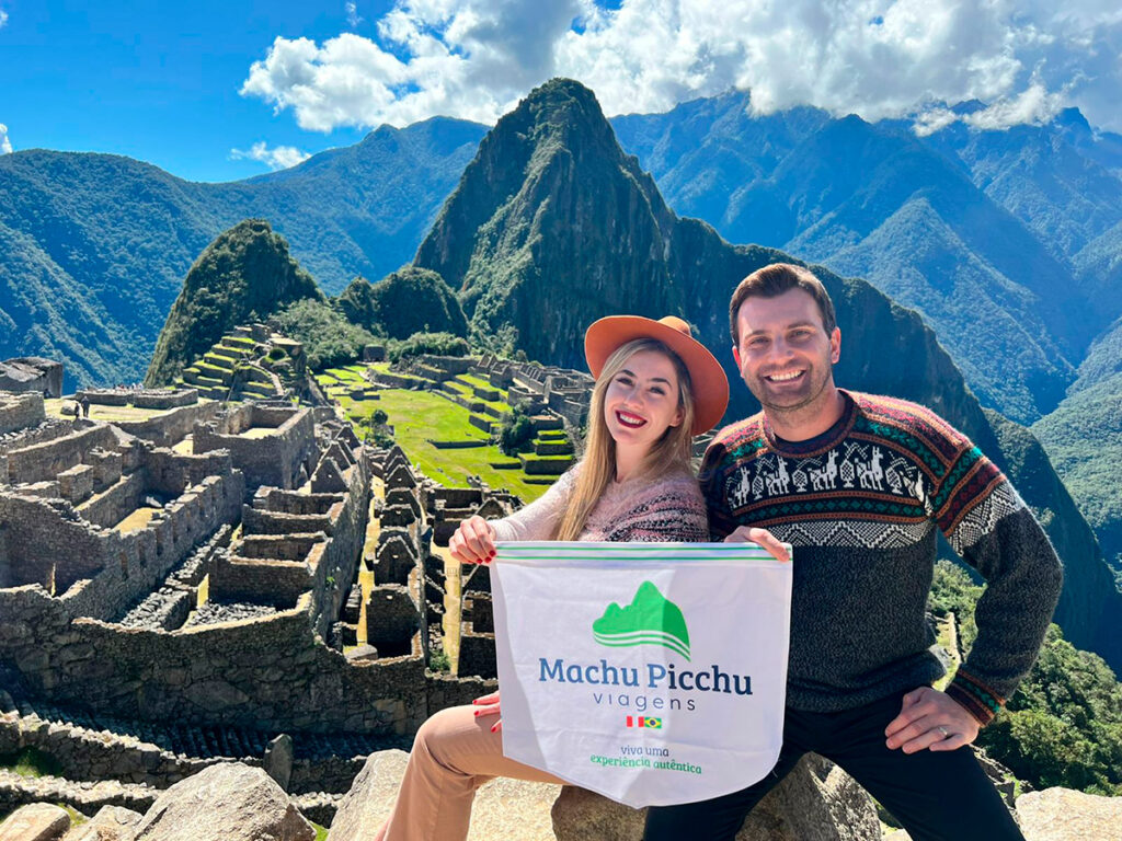 Pareja de viajeros en Machu Picchu con bandera de Viagens Machu Picchu, disfrutando de la vista de la ciudadela inca