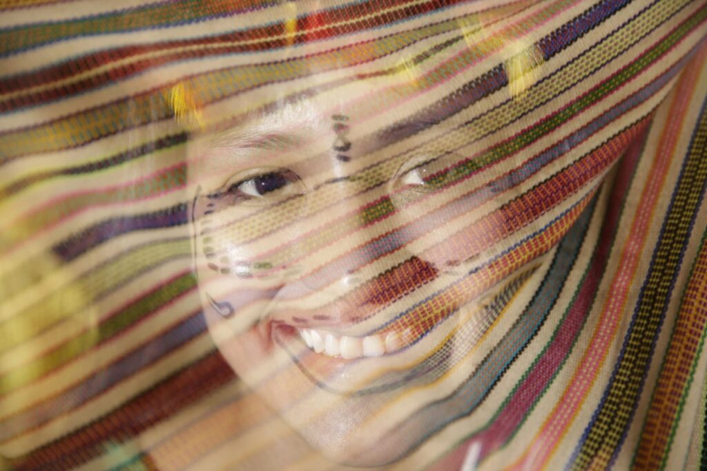 Mujer Yagua con el rostro pintado, vista a través de una tela teñida con colores tradicionales amazónicos