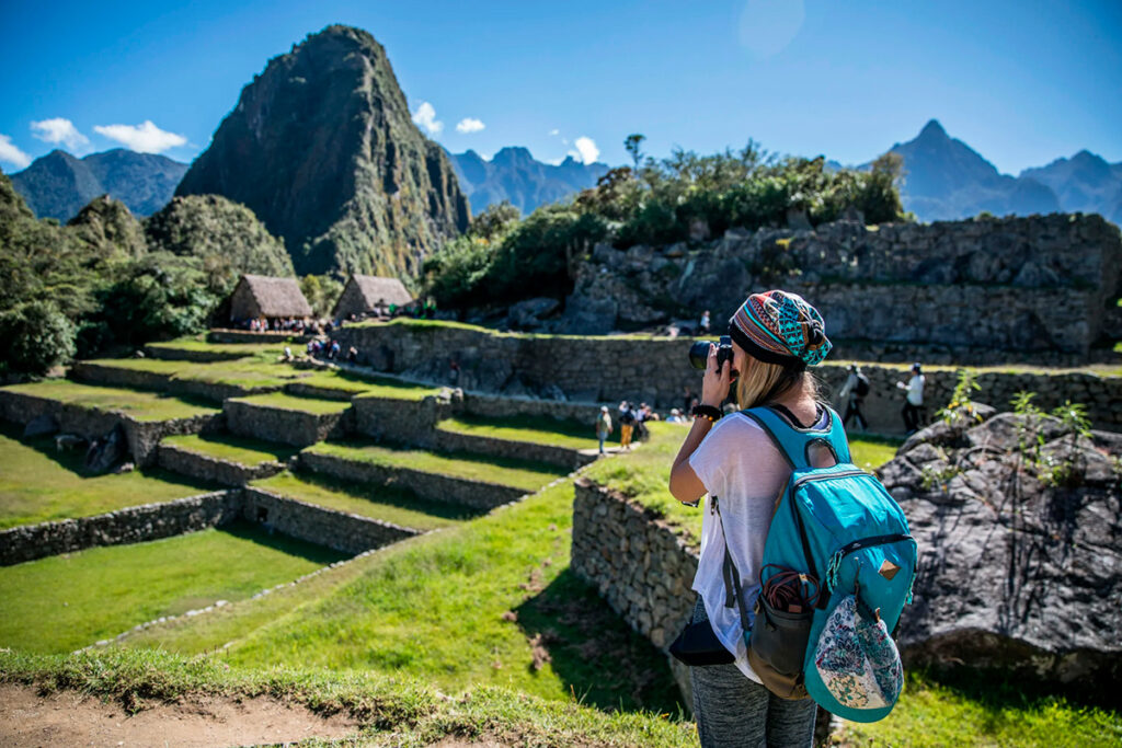 Turista fotografiando Machu Picchu durante un día soleado, rodeada de terrazas y montañas andinas