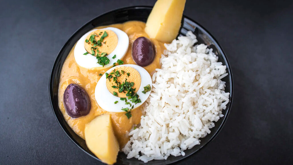 Plato de ají de gallina acompañado de arroz, papas cocidas, huevo duro y aceitunas sobre fondo oscuro