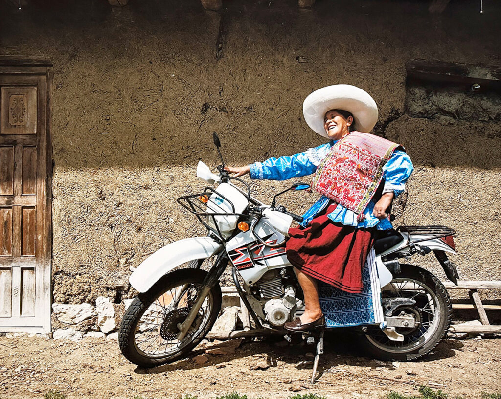 Mujer chotana sonriente, vestida con ropa tradicional y alforja bordada, poosando sobre una motocicleta en un entorno rural