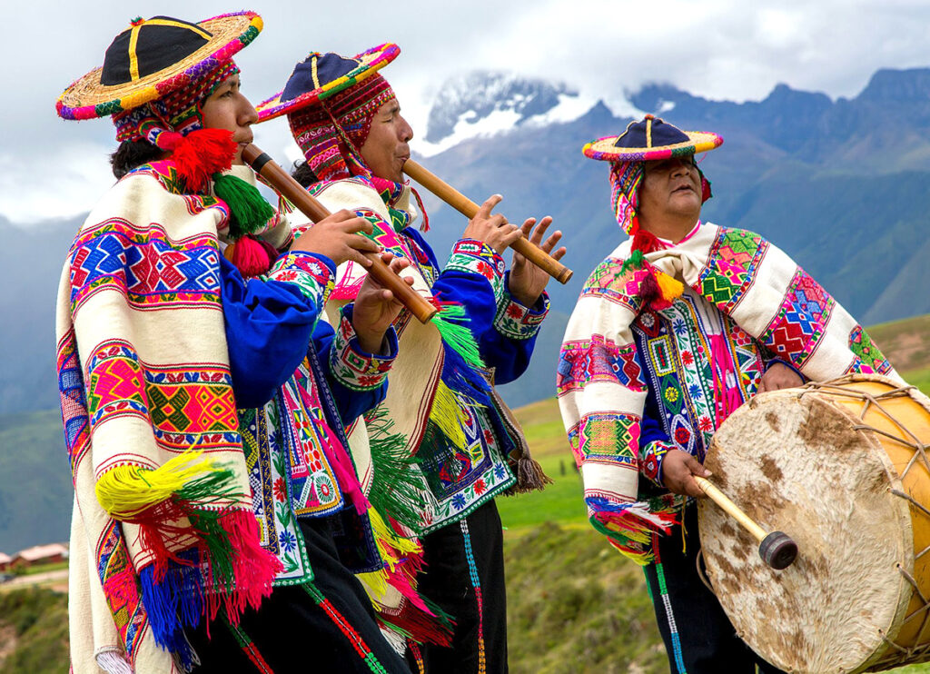 Músicos andinos tocando sikus y percusión durante una festividad tradicional, vestidos con trajes multicolores con el fondo de los andes peruanos