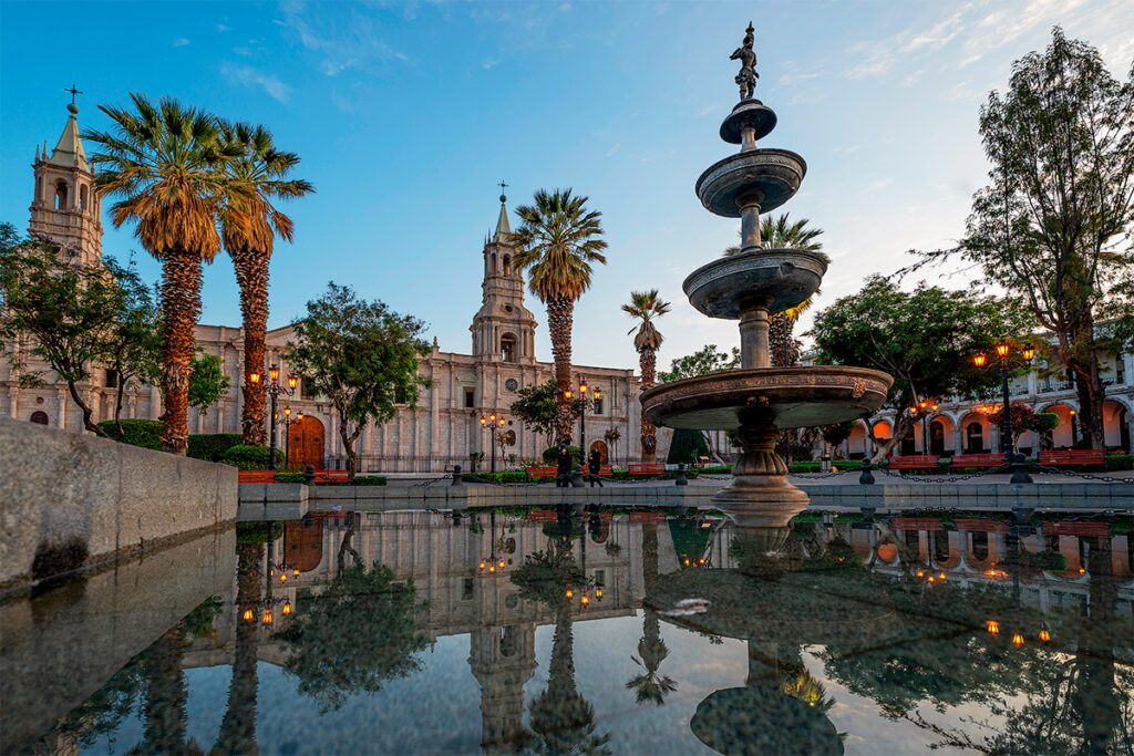 Reflejo de la fuente central y la Catedral de Arequipa al atardecer en la Plaza de Armas, con cielo despejado y palmeras