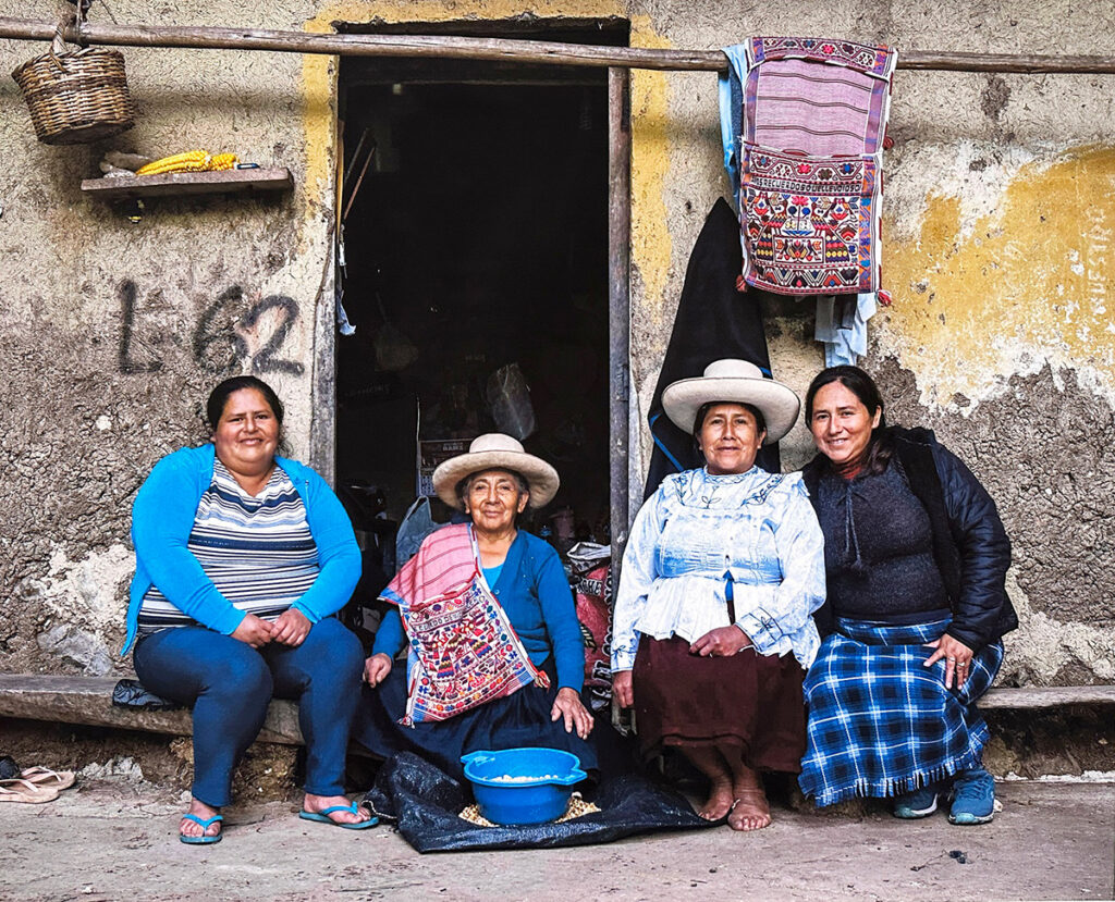 Grupo de mujeres tejedoras chotanas sentadas frente a una vivienda, con alforjas tejidas colgadas y vestimenta tradicional