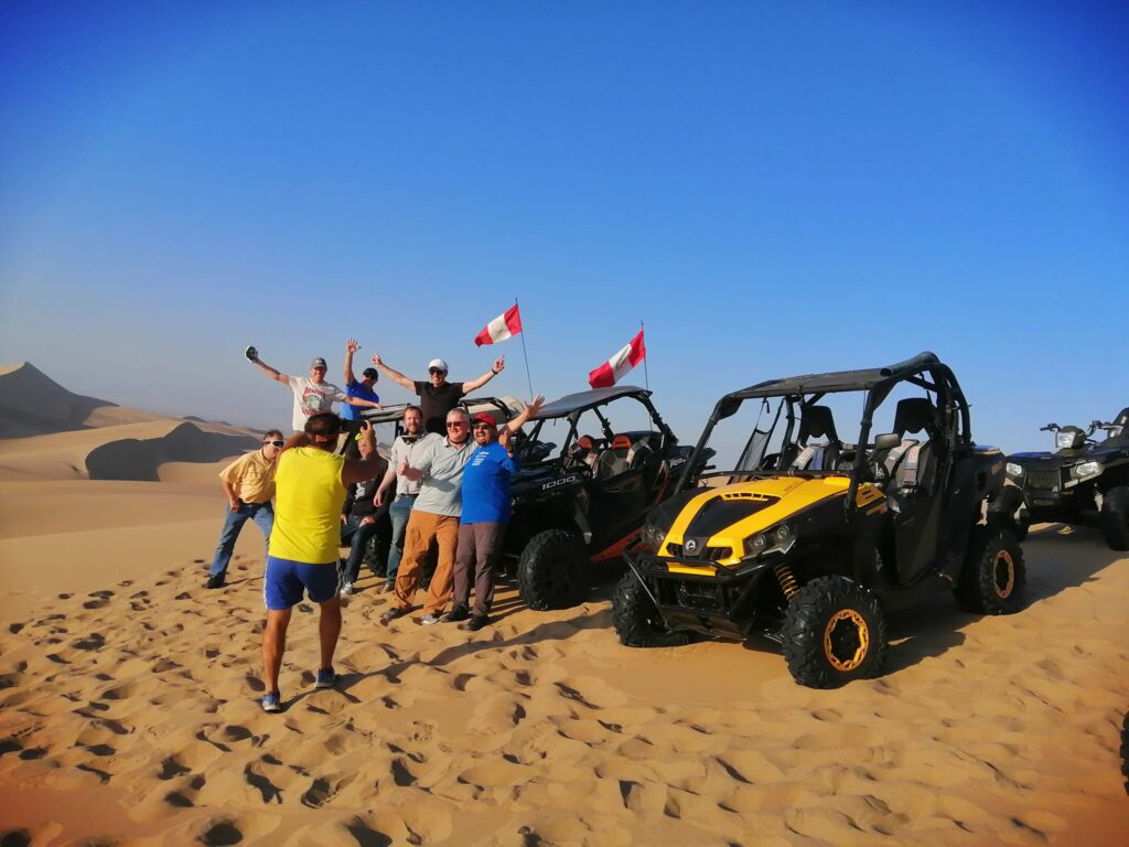Grupo de viajeros en el desierto de Huacachina junto a cuatrimotos y buggies, rodeados de dunas bajo cielo despejado