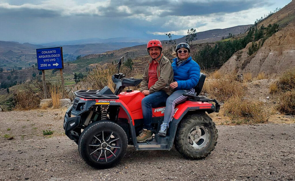 Pareja en cuatrimoto en el borde del Cañón del Colca, con fondo montañoso y señal de sitio arqueológico