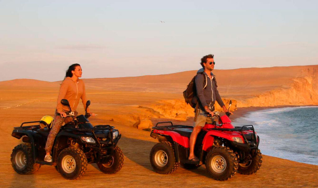 Pareja en cuatrimoto frente al mar en la Reserva Nacional de Paracas, con acantilados y desierto iluminados por la luz del atardecer