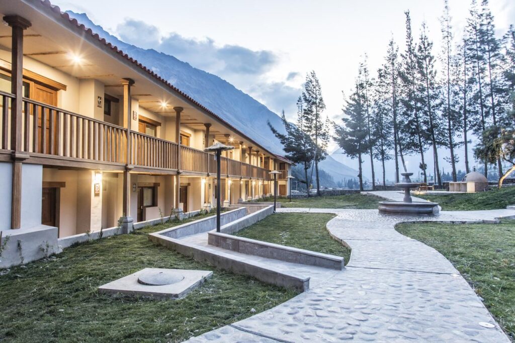 Fachada iluminada del Del Pilar Ollantaytambo Hotel al atardecer, rodeada de árboles y montañas en un entorno rural del Valle Sagrado