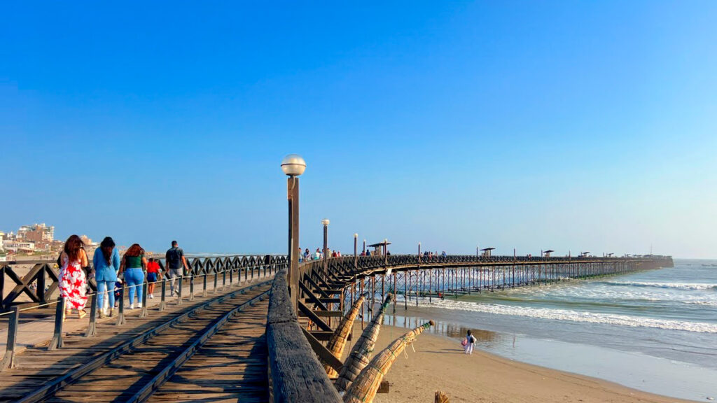Turistas caminan por el muelle de madera de Pimentel en un día soleado, con vista al mar y la playa de Lambayeque, Perú
