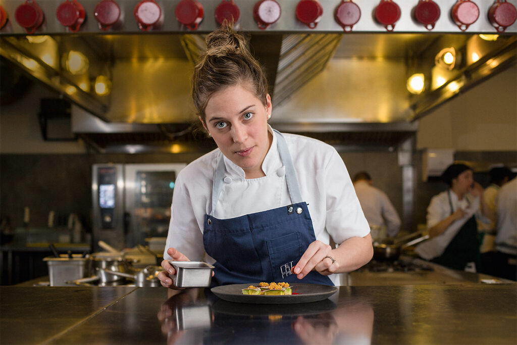 Pía León en la cocina de Kjolle, emplatando uno de sus platos con precisión, frente a un fondo de cocina abierta y equipo en acción