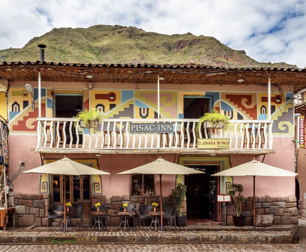 Fachada colorida del hotel Pisac Inn, con balcón adornado y mesas al aire libre frente a la plaza principal del pueblo de Pisac