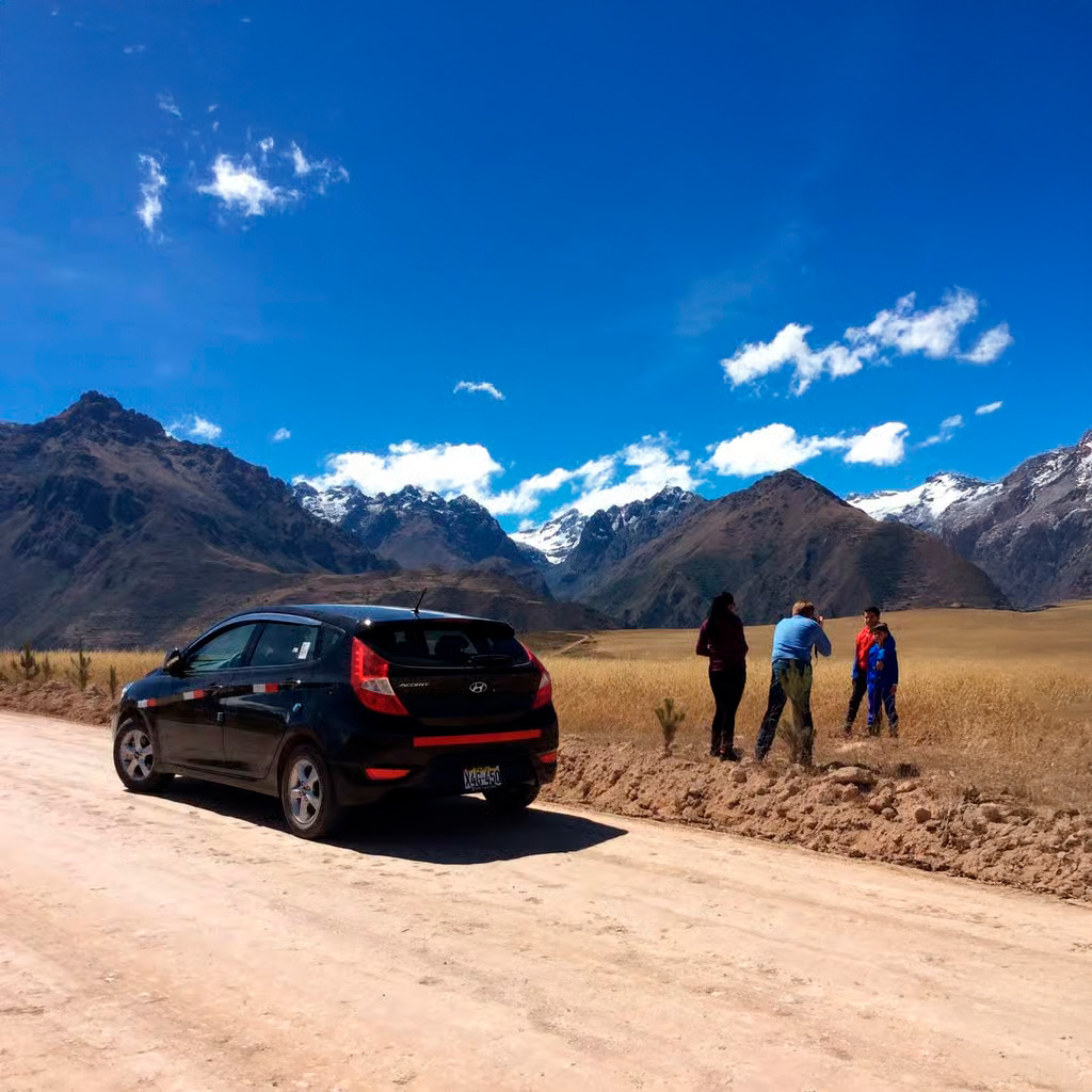 Turistas y automóvil en ruta hacia Moray, con el paisaje montañoso del Valle Sagrado al fondo, en un día despejado