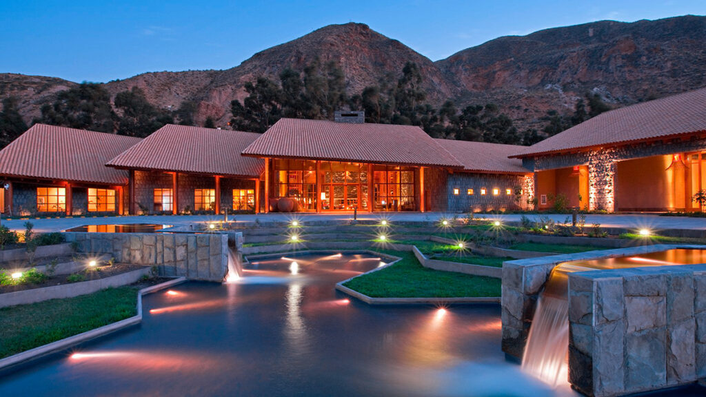 Fachada iluminada del hotel Tambo del Inka al atardecer, con canales de agua y montañas andinas al fondo en Urubamba