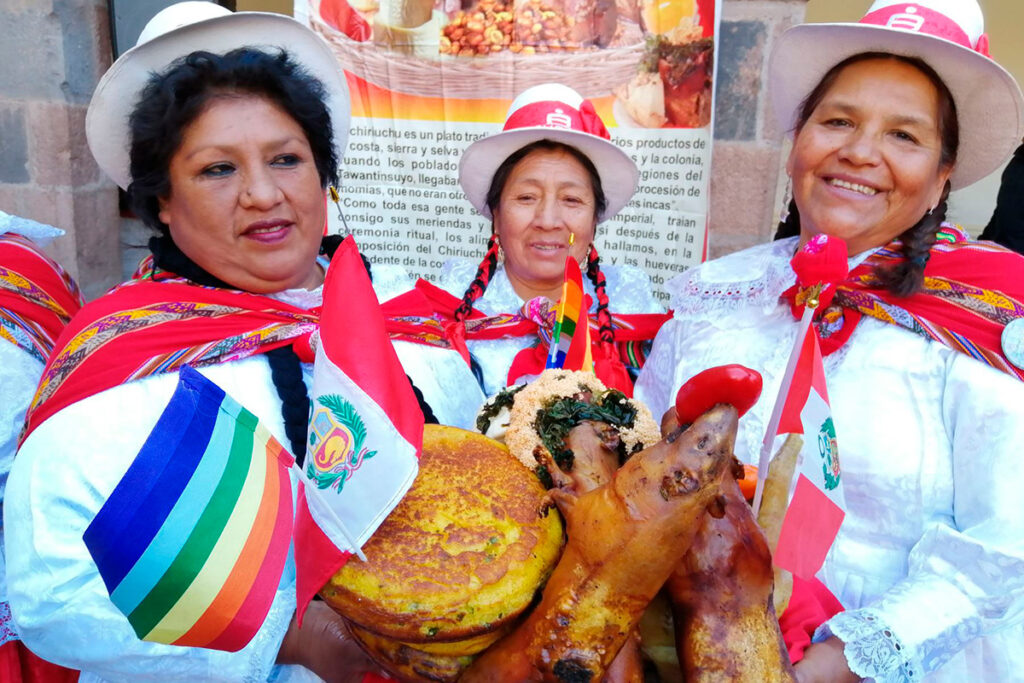 Tres mujeres cusqueñas con vestimenta tradicional sostienen un plato de chiriuchu decorado con banderas del Perú y la wiphala durante el Corpus Christi