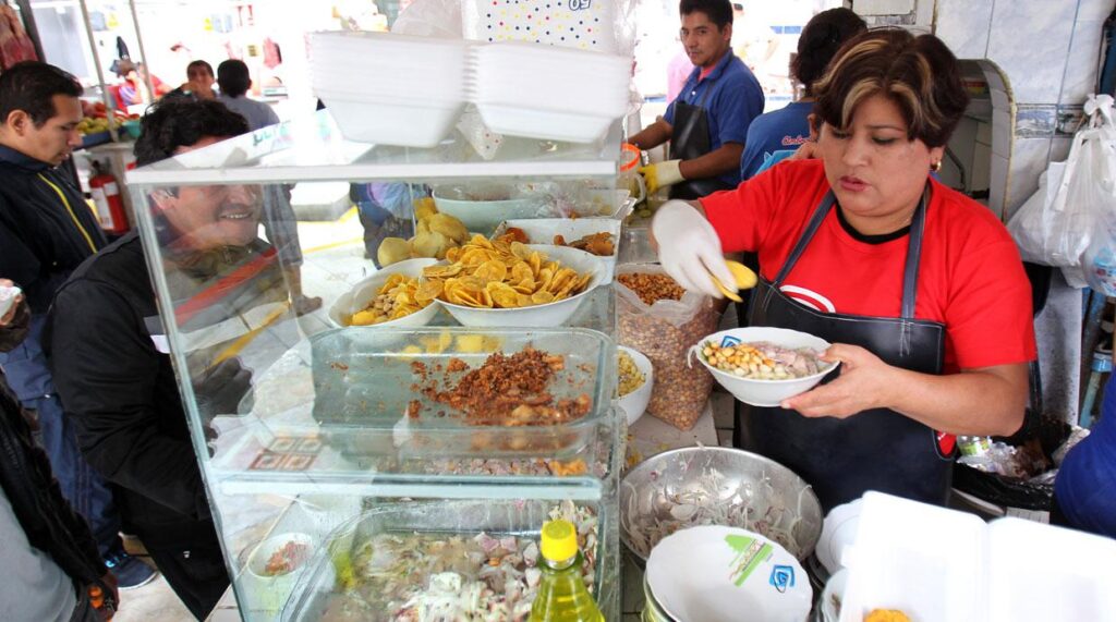 Mujer sirviendo ceviche en un puesto de mercado popular, rodeada de ingredientes tradicionales como camote, cancha, y chifles