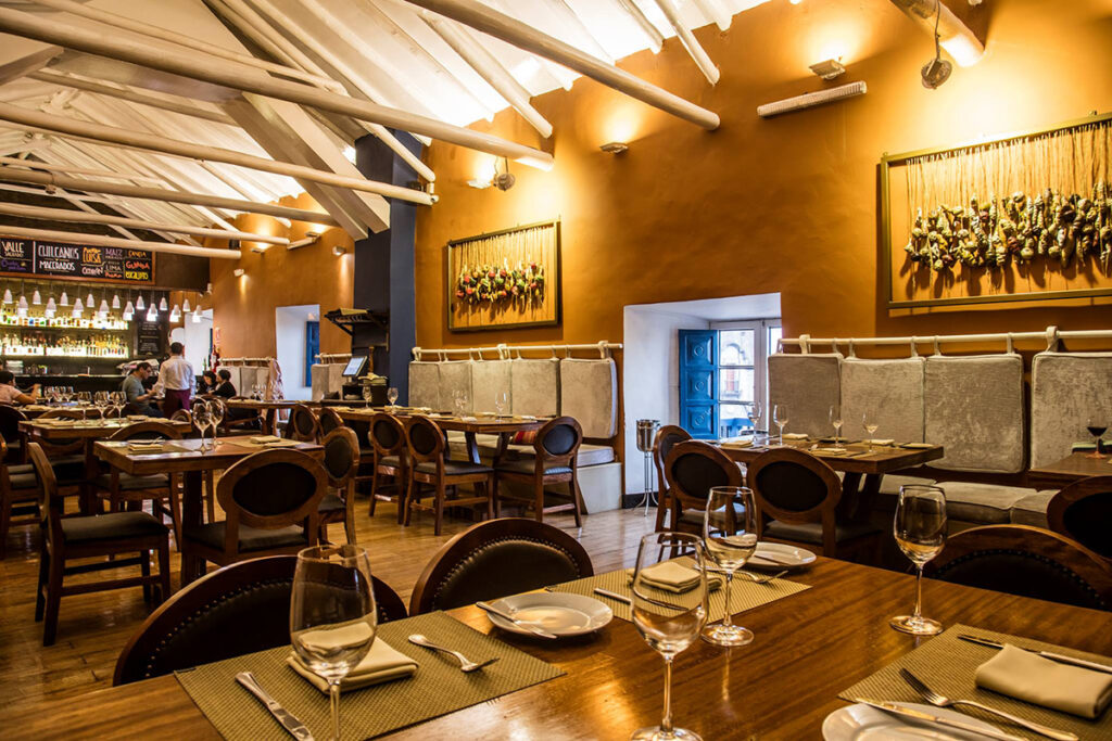 Interior del restaurante Chicha por Gastón Acurio en Cusco, con mesas elegantemente dispuestas, techos altos, decoración cálida y un bar al fondo