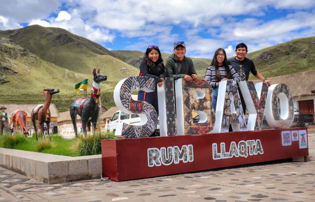 Grupo de jóvenes turistas posando junto al letrero de Sibayo con esculturas de llamas al fondo