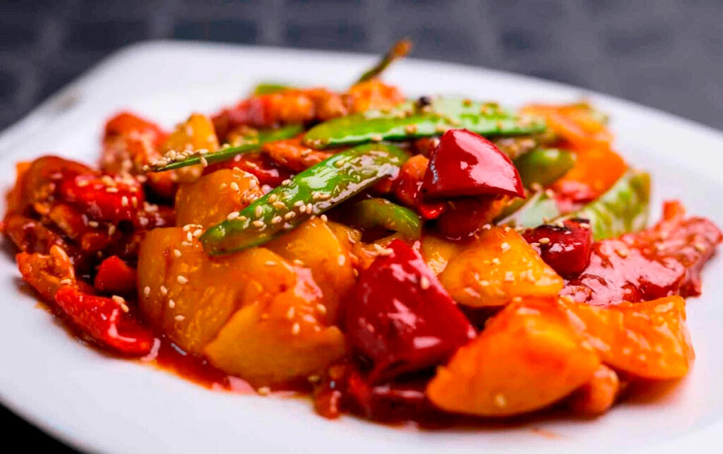 Kam Lu Wantán servido en plato blanco, con trozos de piña, pimientos, vainitas y salsa agridulce