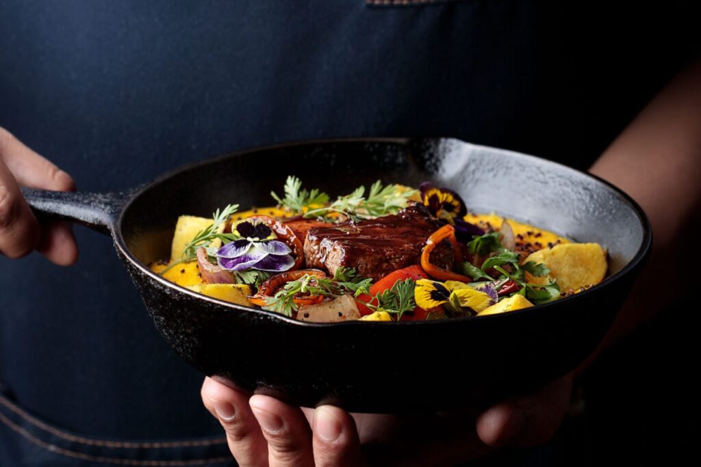 Plato principal servido en Morena Peruvian Kitchen, presentado en sartén de hierro, con carne, vegetales, flores comestibles y guarnición de colores vibrantes