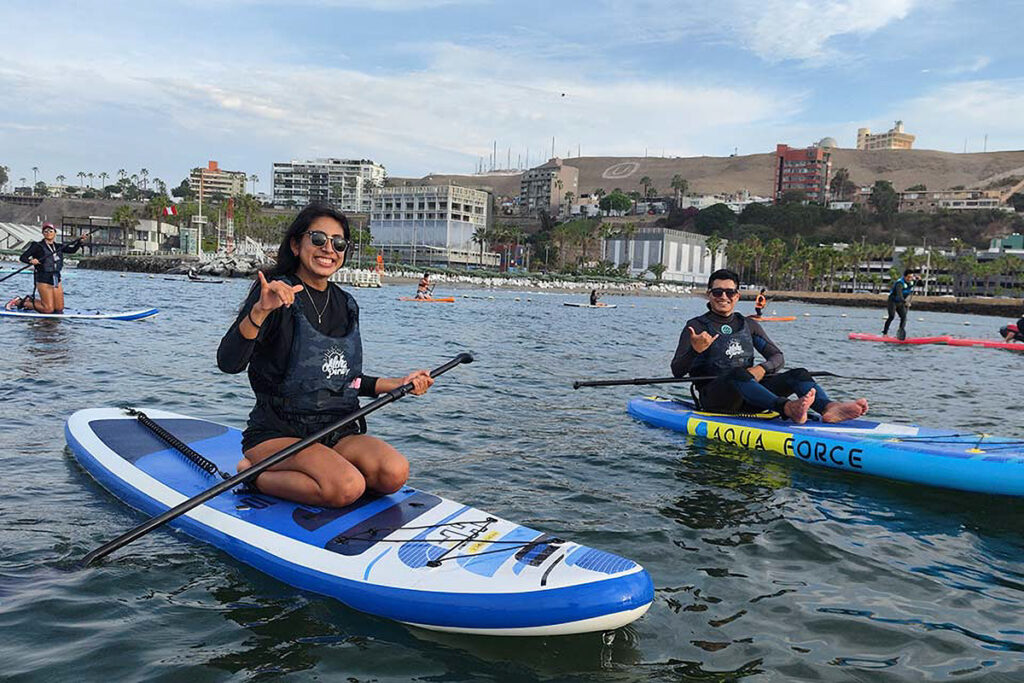 Personas practicando paddle surf en Playa Pescadores, Costa Verde de Lima, con edificios urbanos al fondo