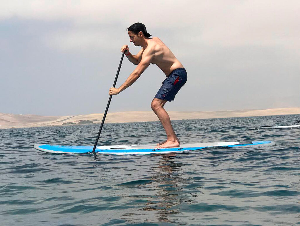 Persona practicando paddle surf en la bahía de Paracas, Perú, con paisaje desértico al fondo.