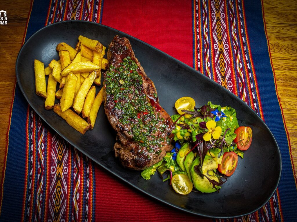 Plato gourmet en Senzo, con corte de carne acompañado de papas fritas, ensalada fresca con flores comestibles, servido sobre mantel tradicional andino