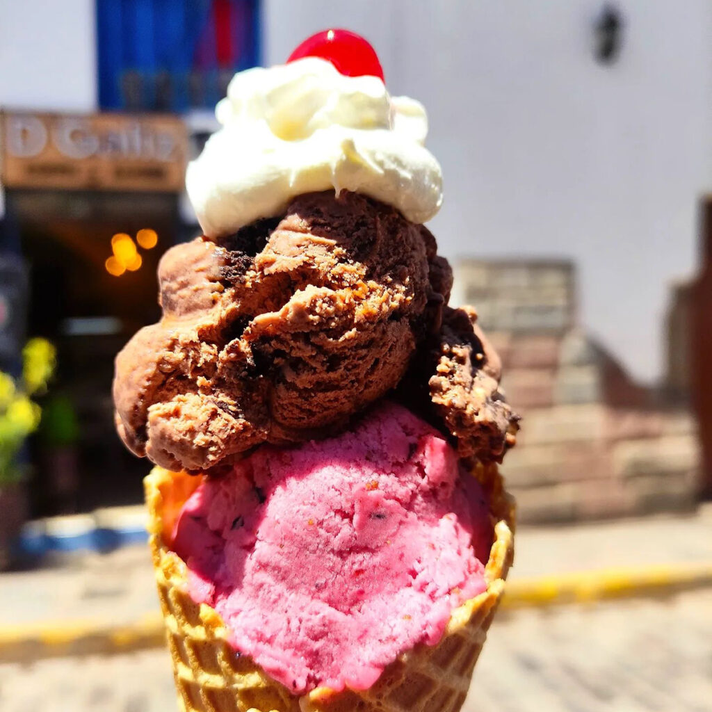 Cono de helado artesanal con sabores rosa y chocolate, decorado con crema batida y cereza, en una calle de Cusco