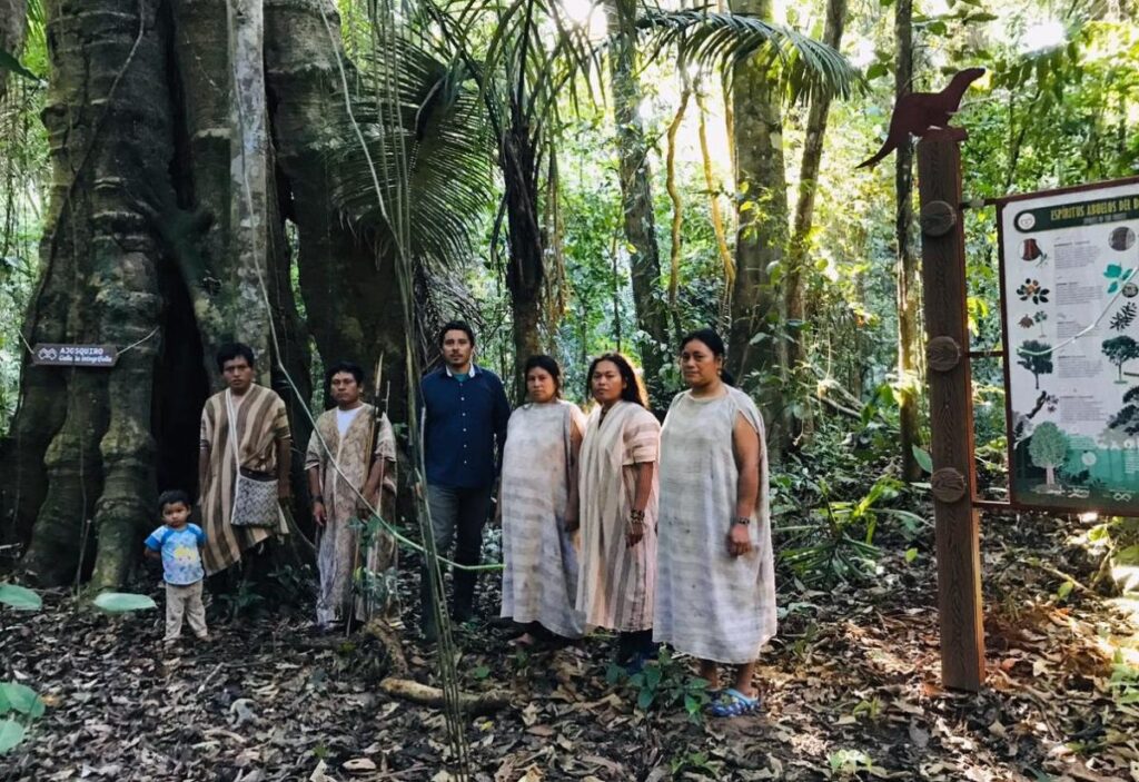 Miembros de una comunidad machiguenga posan junto a un visitante en medio del bosque, cerca de un cartel informativo sobre especies locales