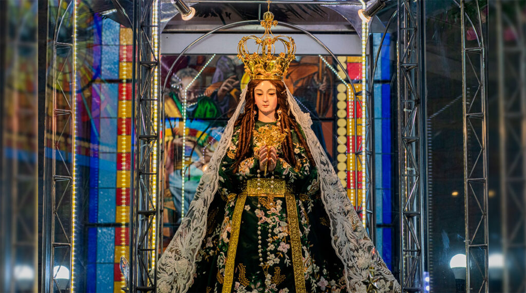 Imagen de la Virgen de la Puerta de Otuzco vestida con manto bordado, corona dorada y manos en posición de oración, dentro de su santuario iluminado