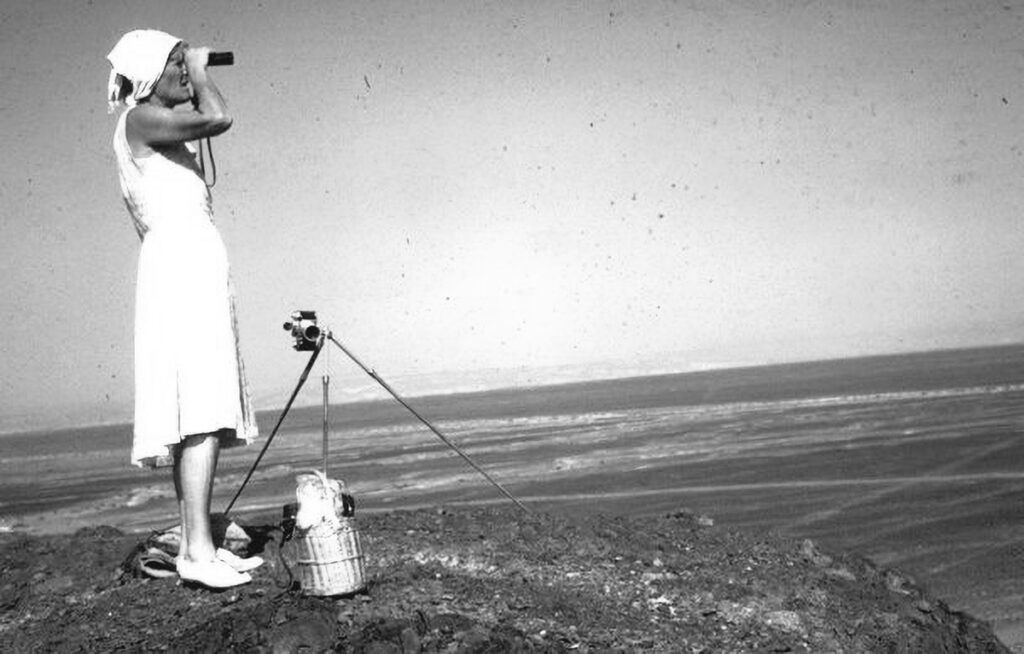María Reiche observando desde un punto elevado en el desierto de Nazca, con binoculares en mano y equipo fotográfico a su lado