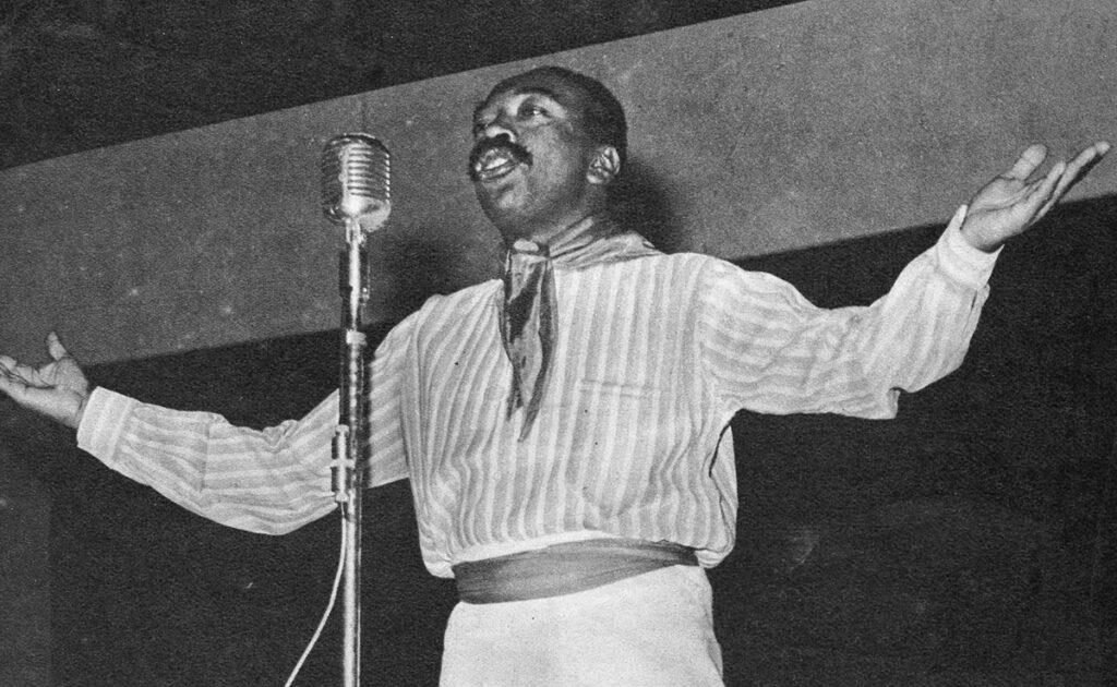 Nicomedes Santa Cruz recitando una décima frente a un micrófono, con los brazos abiertos, en una presentación pública en blanco y negro