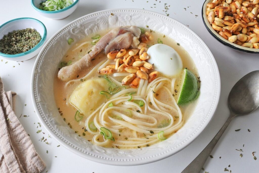 Plato hondo con caldo de gallina, pierna de pollo, huevo duro, papa amarilla y tallarines gruesos, acompañado de maíz tostado y rodaja de limón al costado