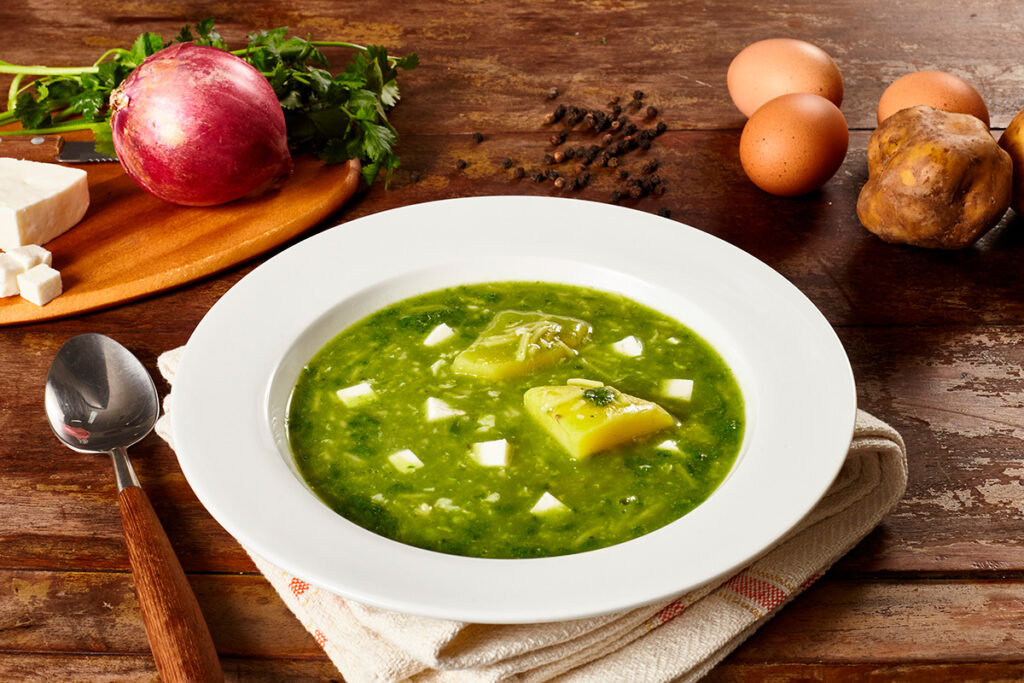 Sopa tradicional del norte con papa amarilla en trozos, queso fresco y huevo, todo sumergido en caldo verde de hierbas aromáticas, presentada en plato hondo blanco sobre mesa rústica