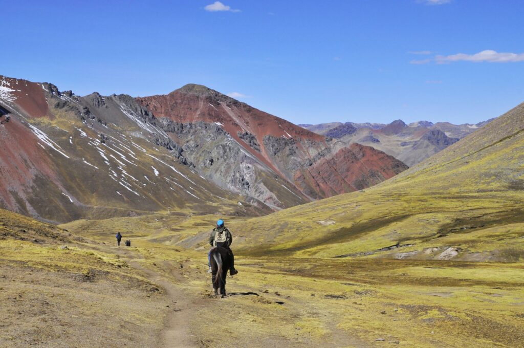 Turista a caballo en el sendero hacia la Montaña Vinicunca, rodeado de montañas de colores y paisajes andinos en Cusco